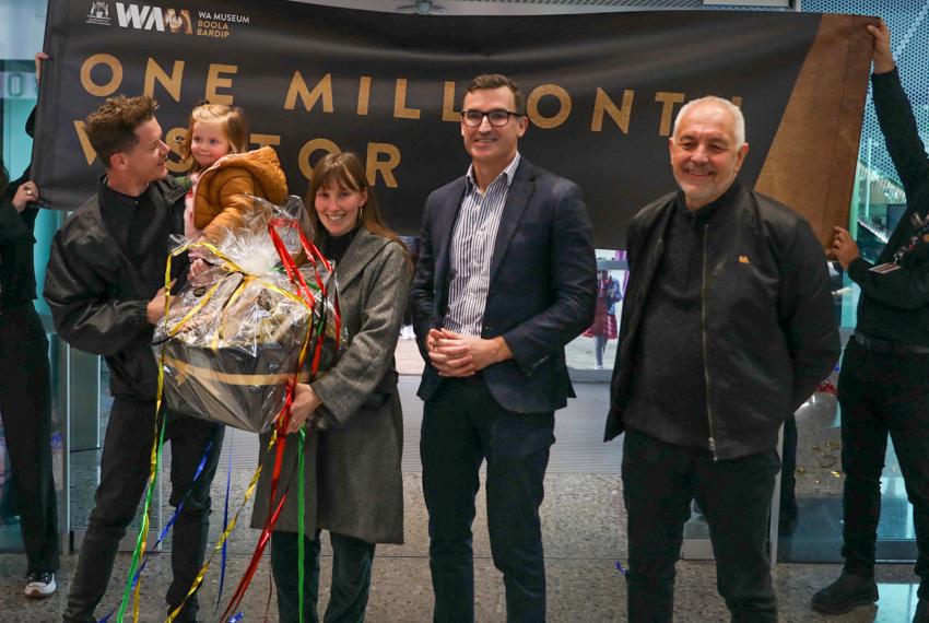 Four adults and a toddler stand in front of a banner that says "One Millionth Visitor" held by two Museum staff members.