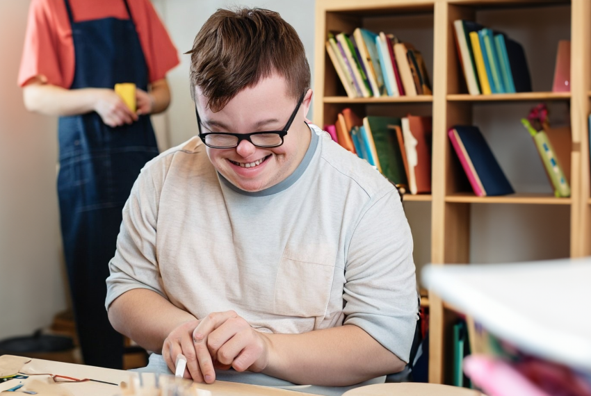 A young man with downs syndrome smiles down at a craft project they're making with string and popsicle sticks