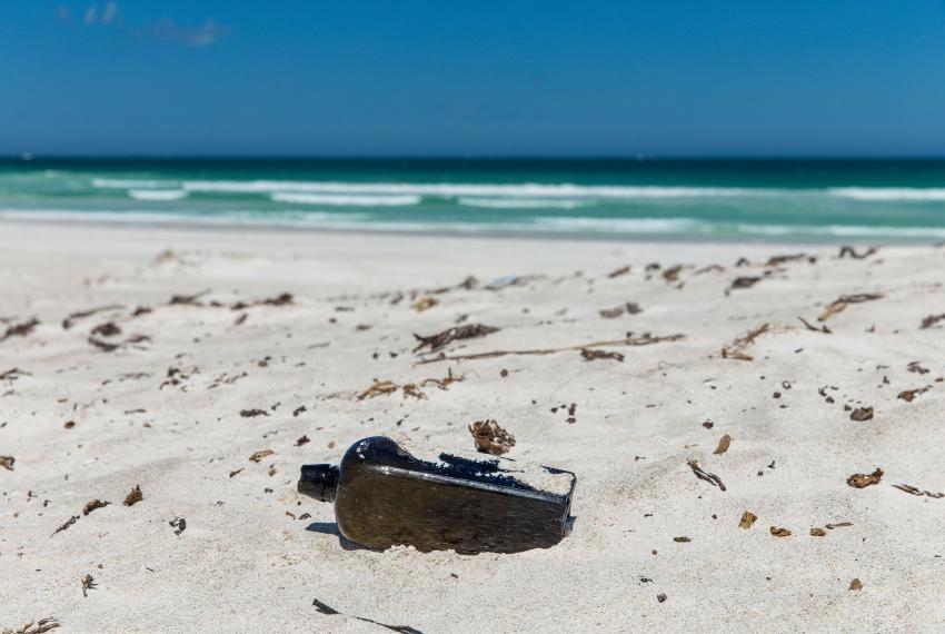 "The bottle is half submerged in sand in the foreground, with crashing waves in the background."