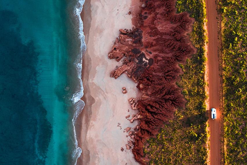 Ocean, red dirt and car driving along a dusty road - aerial shot.
