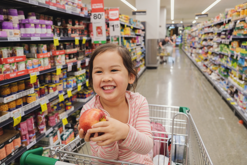  child sitting in a supermarket trolley holds a red apple while shopping in a grocery aisle lined with canned and packaged goods.