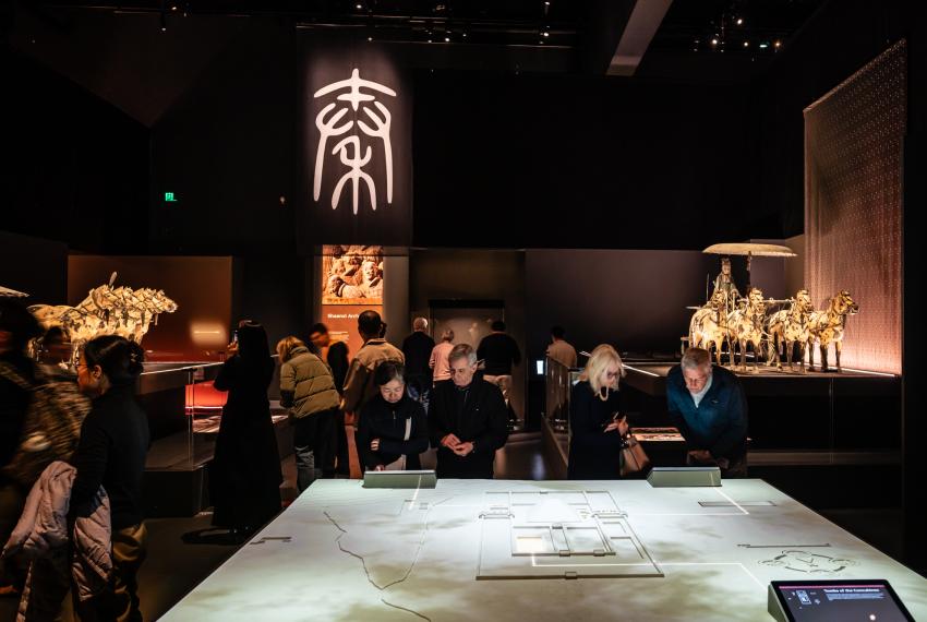 People exploring the Terracotta Warriors exhibition. There are several museum-goers looking at a digital display table with ancient artefacts, including a horse-drawn chariot, in the background.