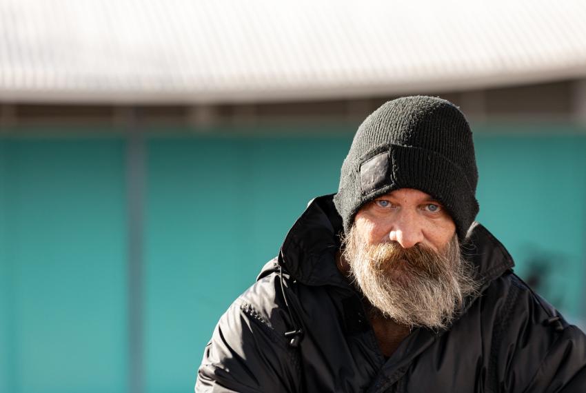 an adult with a beard wearing a beanie stares straight ahead into the camera