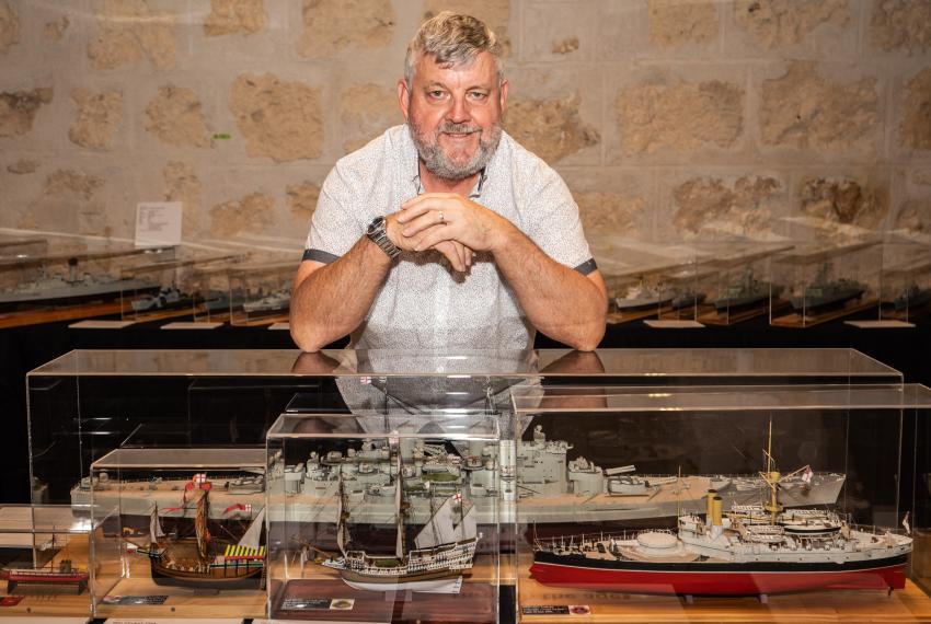 Person sitting behind a display case filled with detailed model ships against a stone wall backdrop.