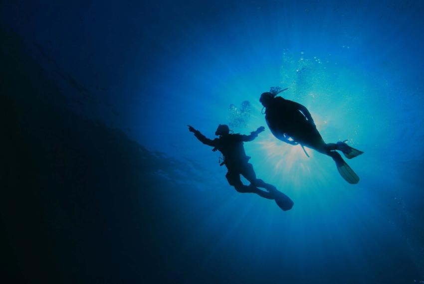 Two scuba divers silhouetted against bright blue sunlight underwater, swimming upward toward the surface.