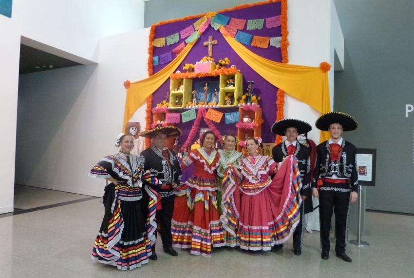 A group of people in colourful traditional costumes standing in front of a decorated Día de los Muertos altar inside a gallery space.