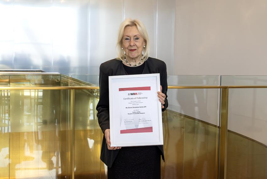 A woman with blonde hair in a black dress holds a certificate up against a gold background