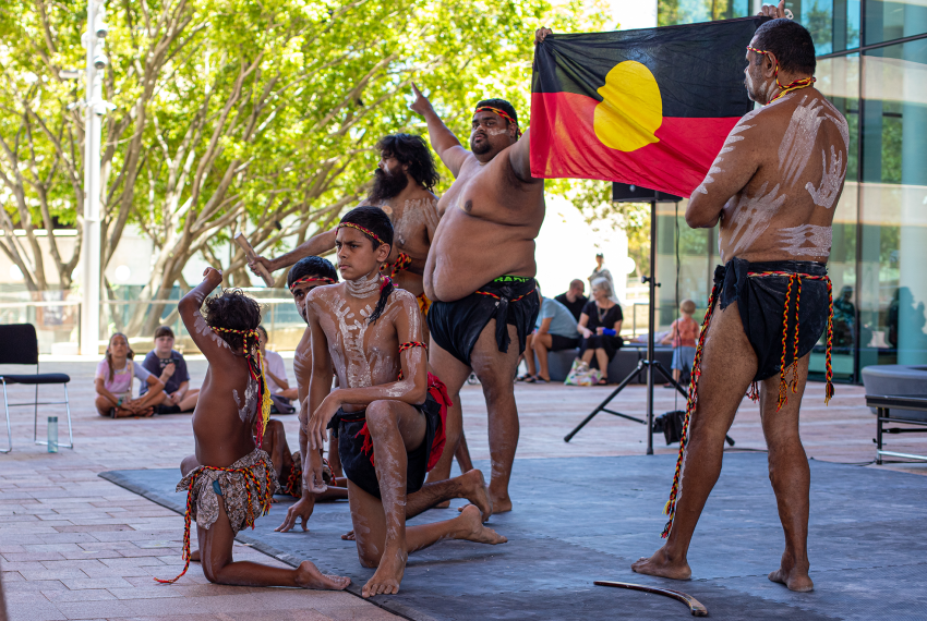 People wearing traditional body paint perform a cultural dance on an outdoor stage, with one person holding an Aboriginal flag while others sit and watch nearby.