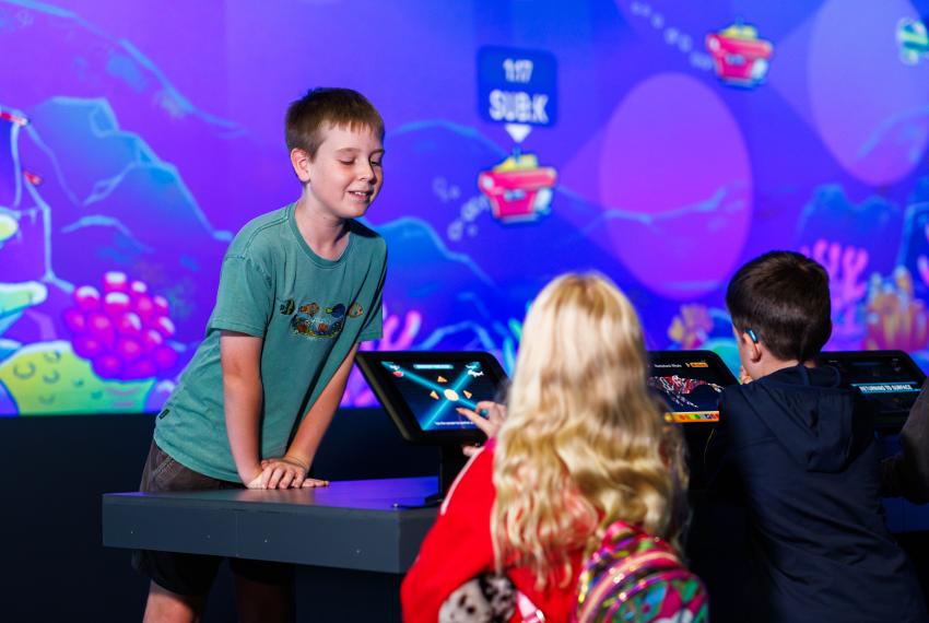 Kid looking over at a girl playing on an ipad with a large screen behind with sea creatures and submarines.