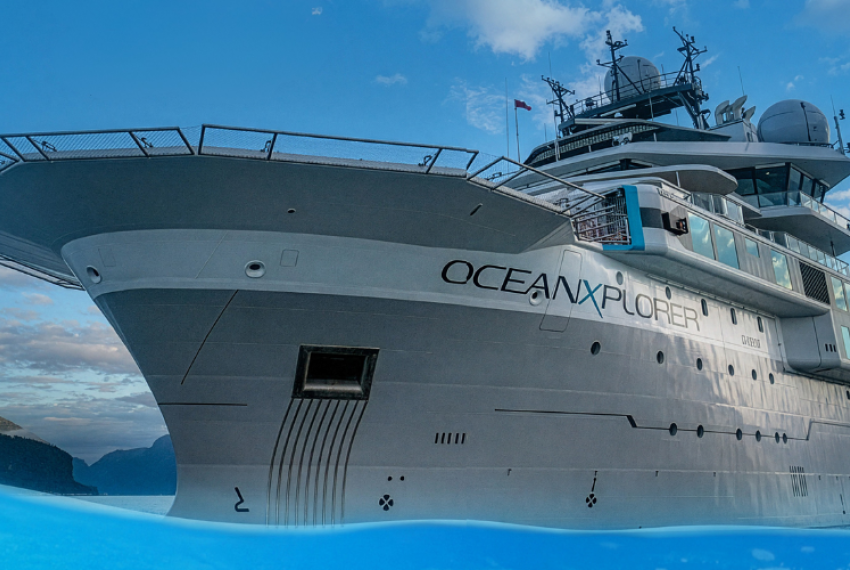 Large white research vessel “OceanXplorer” viewed from a low angle at sea, with blue sky, distant islands, and water partially framing the foreground.