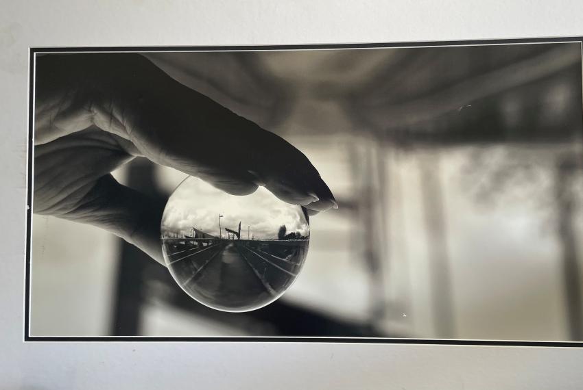 Black-and-white photo of a hand holding a glass sphere that reflects an upside-down view of a pier and cloudy sky.