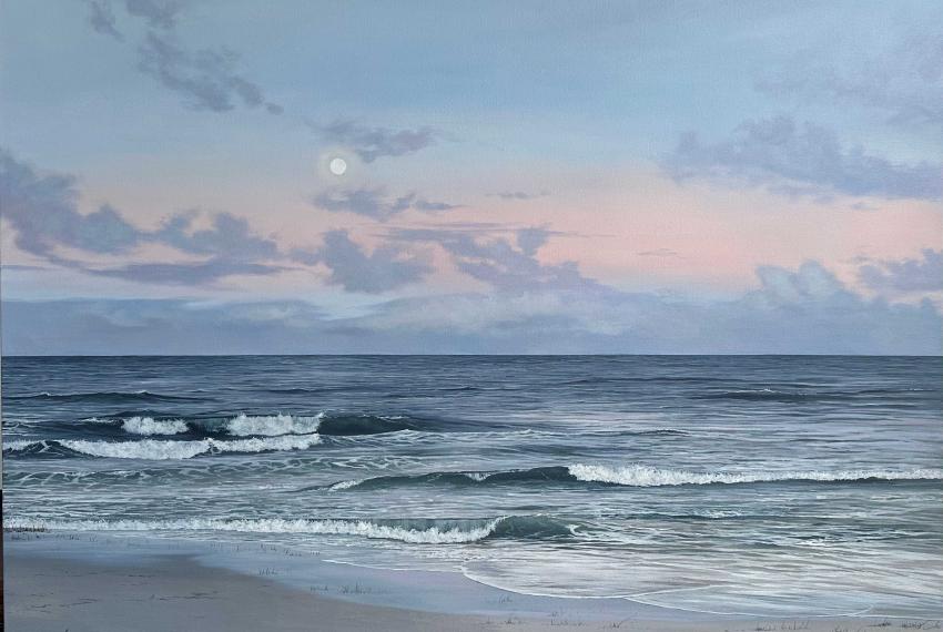 A tranquil seascape showing gentle waves rolling onto a sandy beach beneath a pastel evening sky. Soft pink and blue clouds surround a pale moon above the horizon, reflecting the calm atmosphere of twilight.