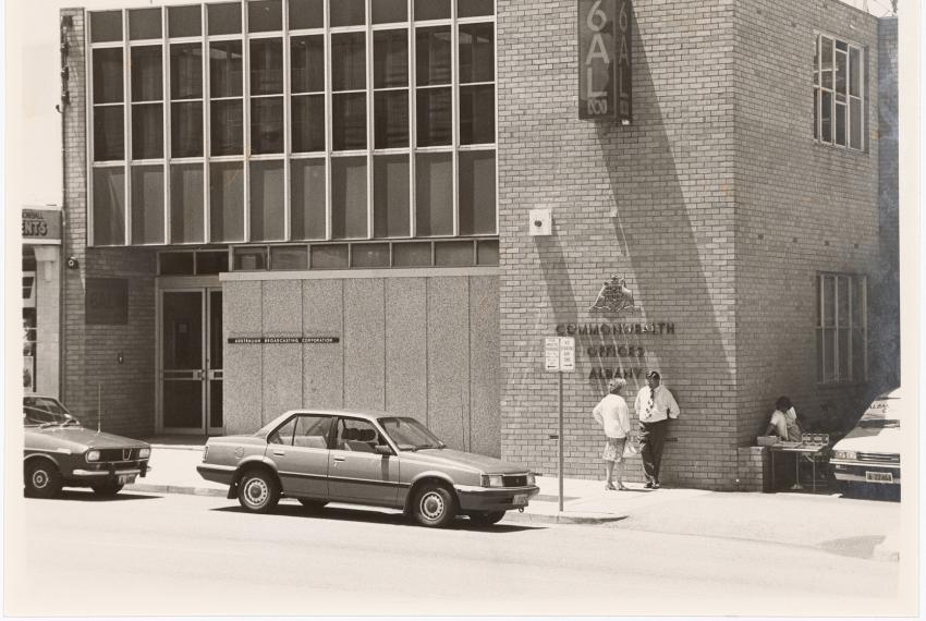a black and white photograph of a building. the building has a sign on it 6AL and another sign Commonwealth Offices Albany. There are cars partked outside the building on the street and a few people standing next to the building