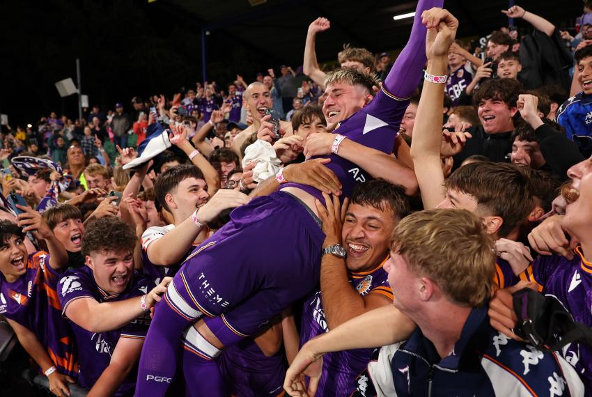 Perth Glory player celebrates with supporters in the stands after a match, surrounded by a cheering crowd in club colours.