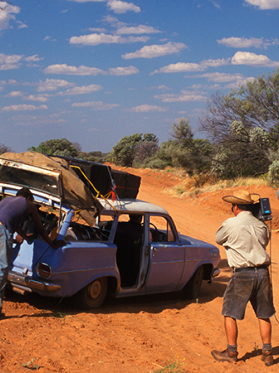 Three people push on an old, blue car bogged on a dirt track, whilst 2 camera people film.