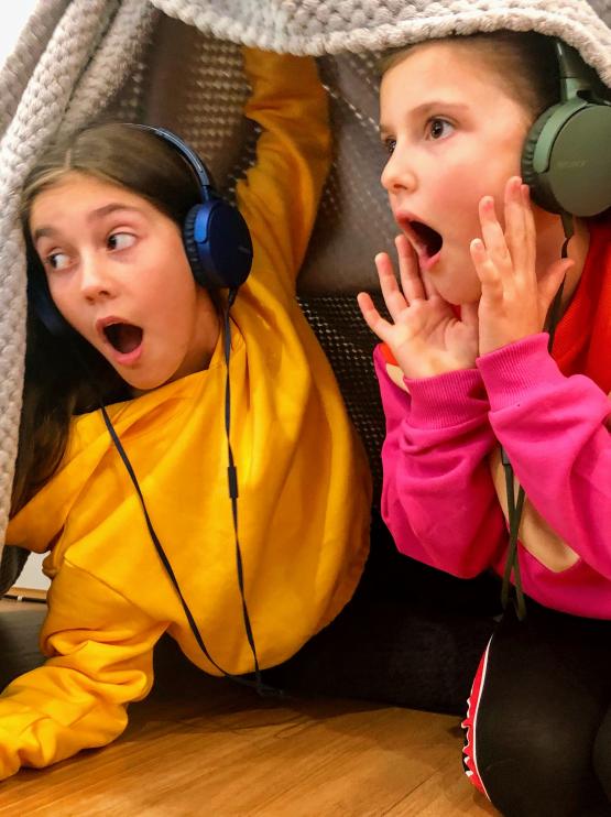 two girls in birght jumpers with headphones on shout and play under a blanket