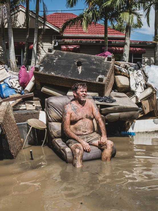 Man sits on a pile of household items washed up in the middle of a flood with a house in the background