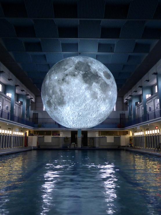 A large model of a moon glows with an etherial white light over a narrow public swimming pool which is backlit in white and yellow, creating a glowing reflection of colour in the otherwise darkened space