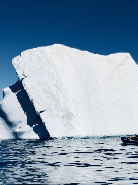 A large white iceberg is framed against a brilliant blue sky and clear rippling blue water, with a tiny boat of researchers at the bottom right corner of the ice berg showing its sheer scale