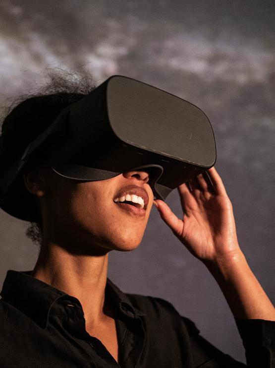 A person wearing a black button up shirt wears a black VR set and smiles as though out of wonder and enjoyment as they look through the headset. They stand in front of a galaxy backdrop