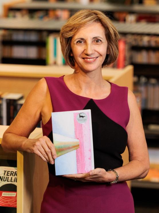 Lisa stands looking at the camera and smiling. She is leaning against a bookshelf and holding her award winning book. She has brown hair with highlights and is wearing pink and black dress. 