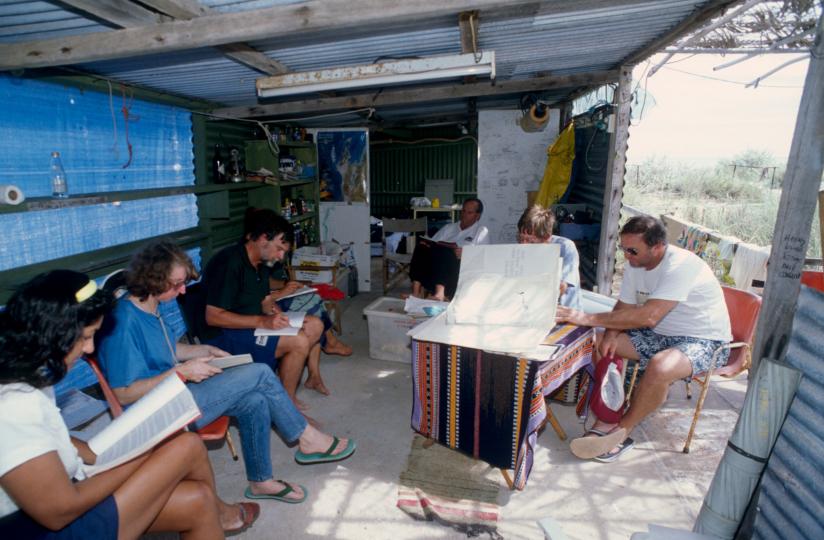 Seven people sitting in a corregated iron hut looking at documents.