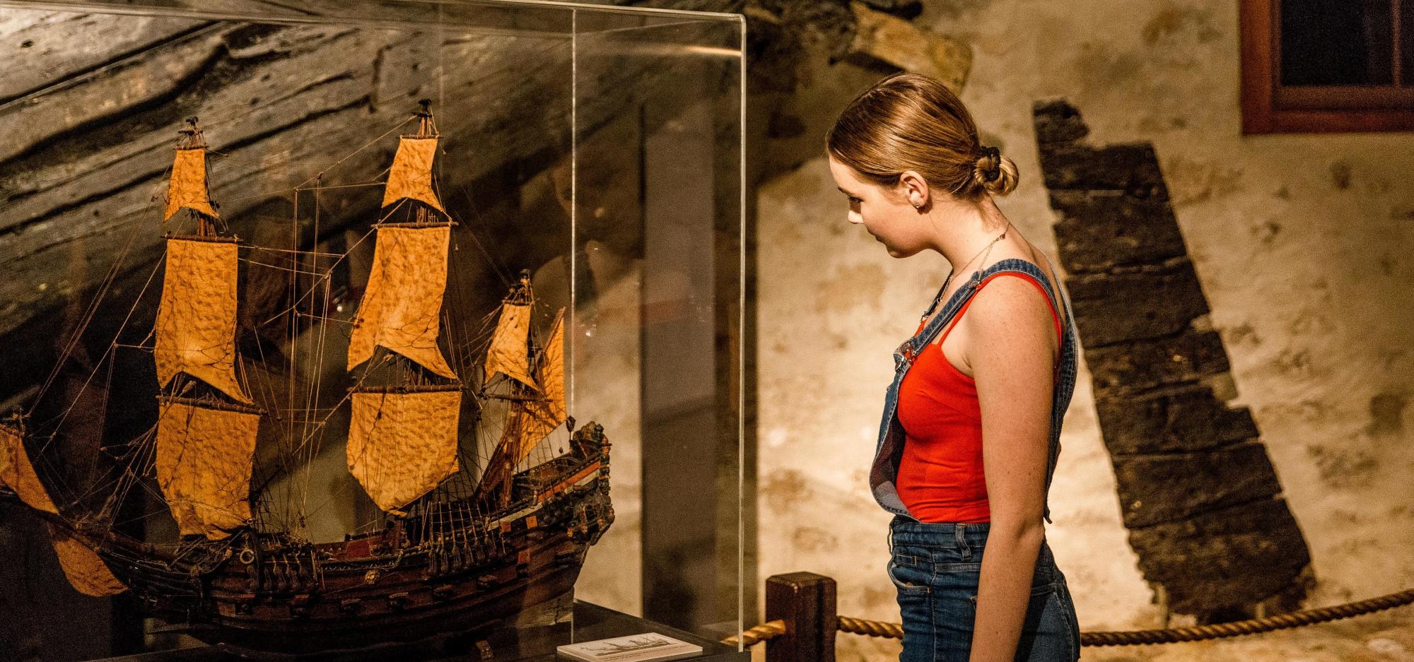 A person stands in a museum gallery, examining a model ship in a showcase