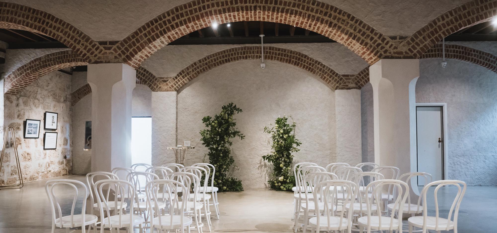 Chairs and flowers are arranged for a wedding in a large room with limestone walls and an arched ceiling