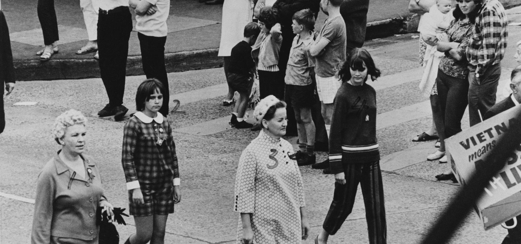 A black and white photograph of people dressed in 1960s-style clothing walking on a street