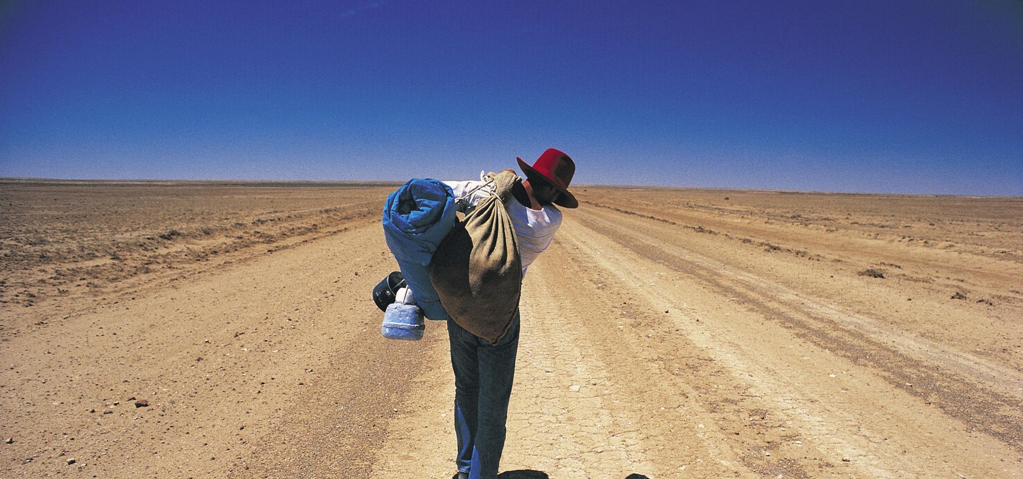 A person wearing a broad-brimmed hat and carrying a swag on their back walks away from the camera along a dusty outback road beneath a clear blue sky