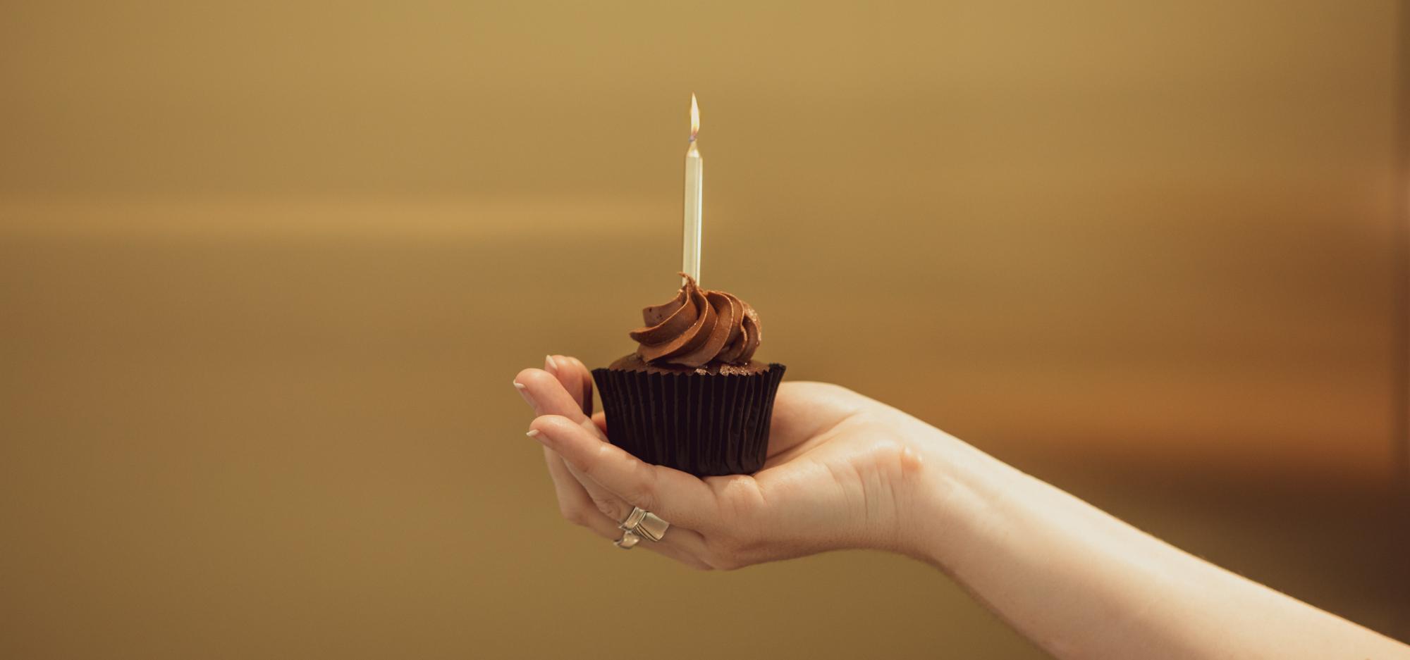 A hand holding a chocolate cupcake with a golden birthday candle