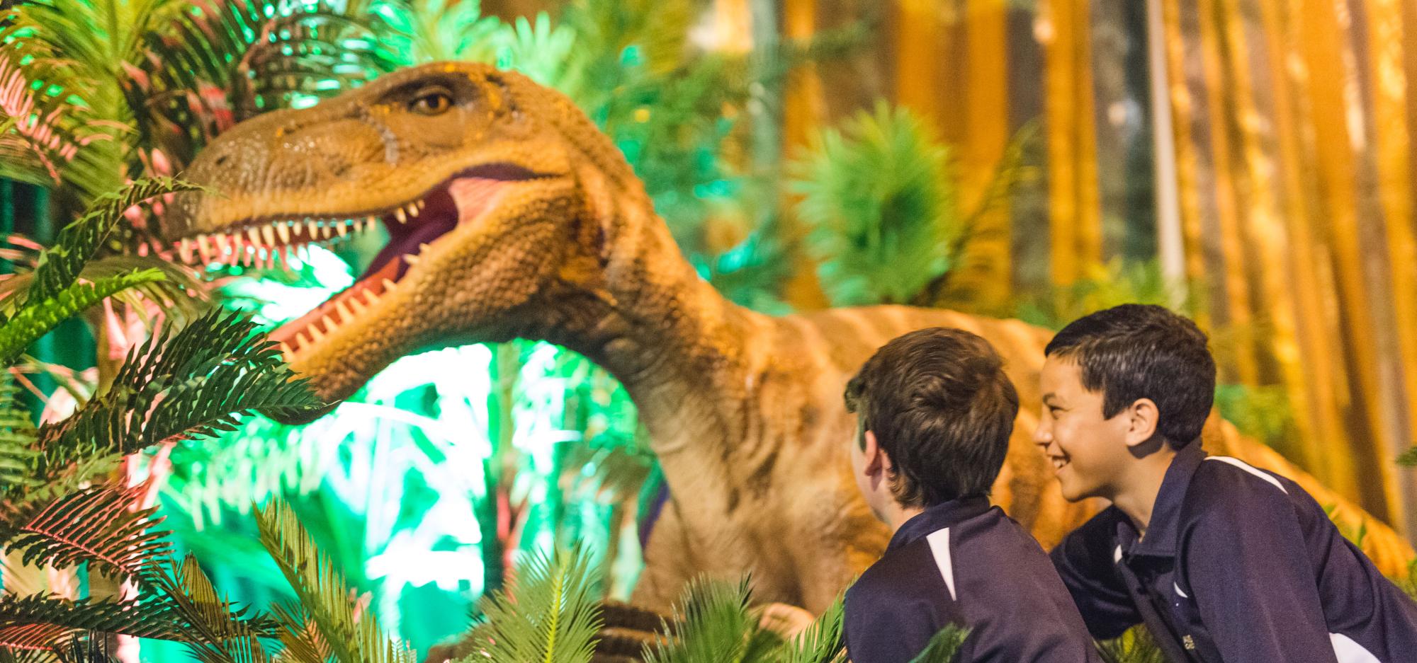 Two children look excitedly at a moving dinosaur puppet standing in a leafy garden