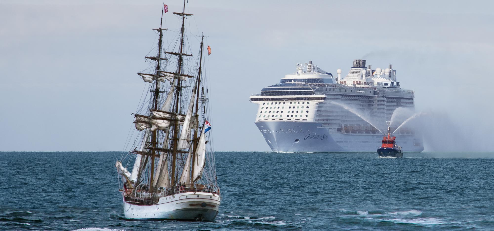 A masted ship sails on the open ocean in front of a modern ocean cruise liner