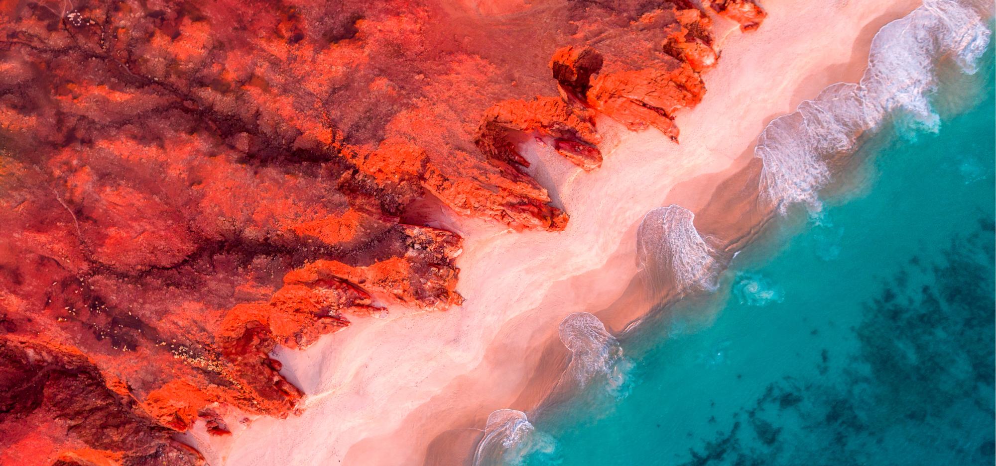 An aerial view of red cliffs beside a clear turquoise sea