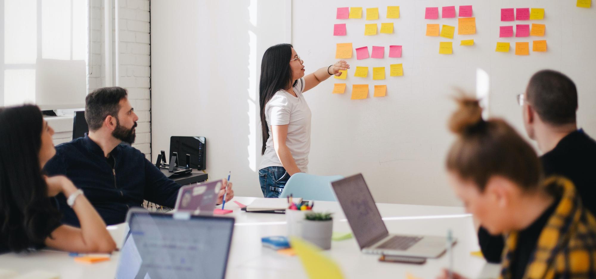 A room full of people sitting at a desk with a person pointing to sticky notes on a whiteboard