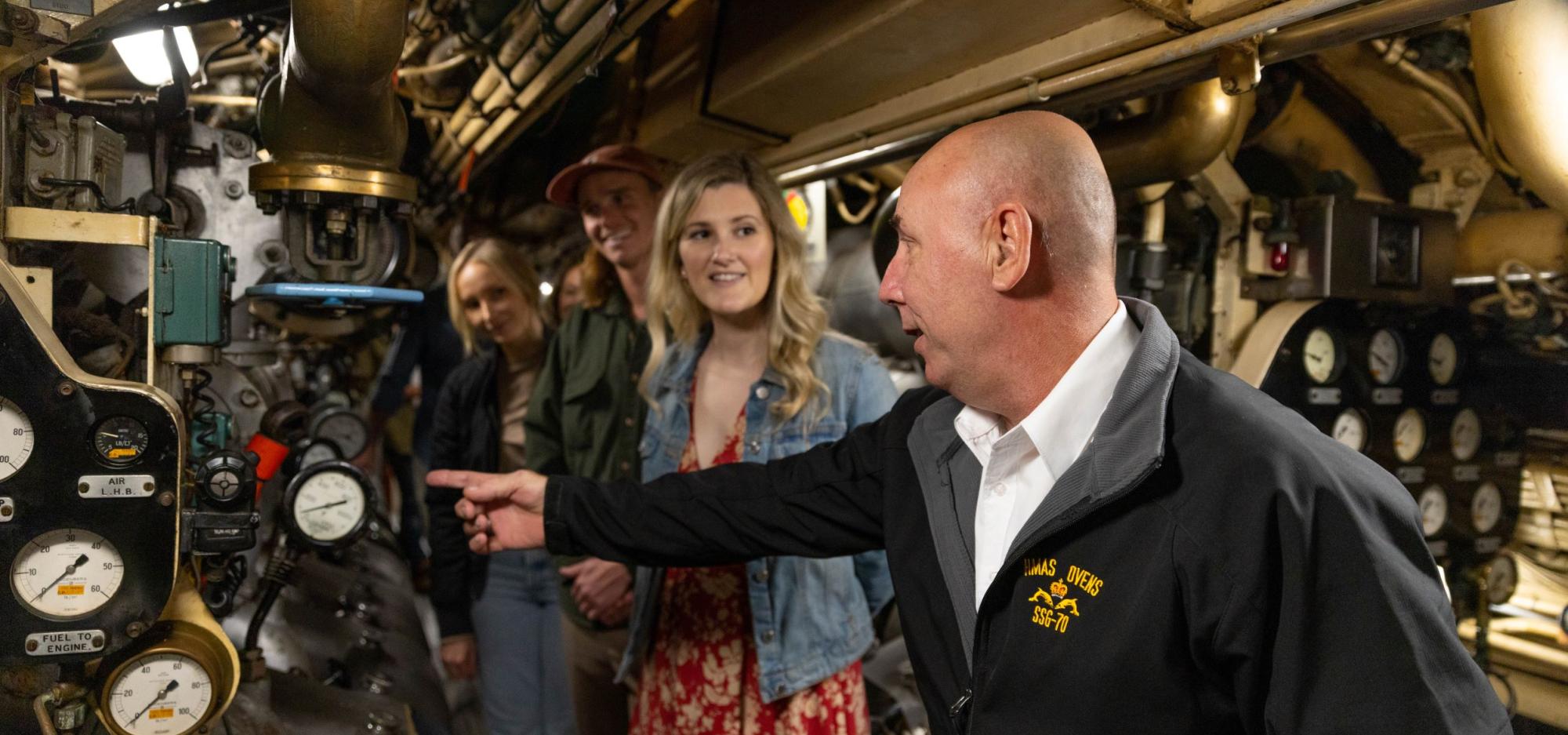 Submarine tour guide gestures inside submarine, three people look on