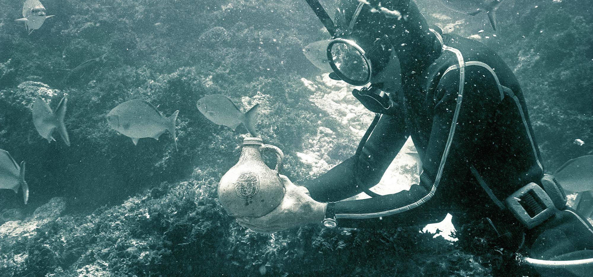 A diver underwater at a shipwreck site examines a pottery jug