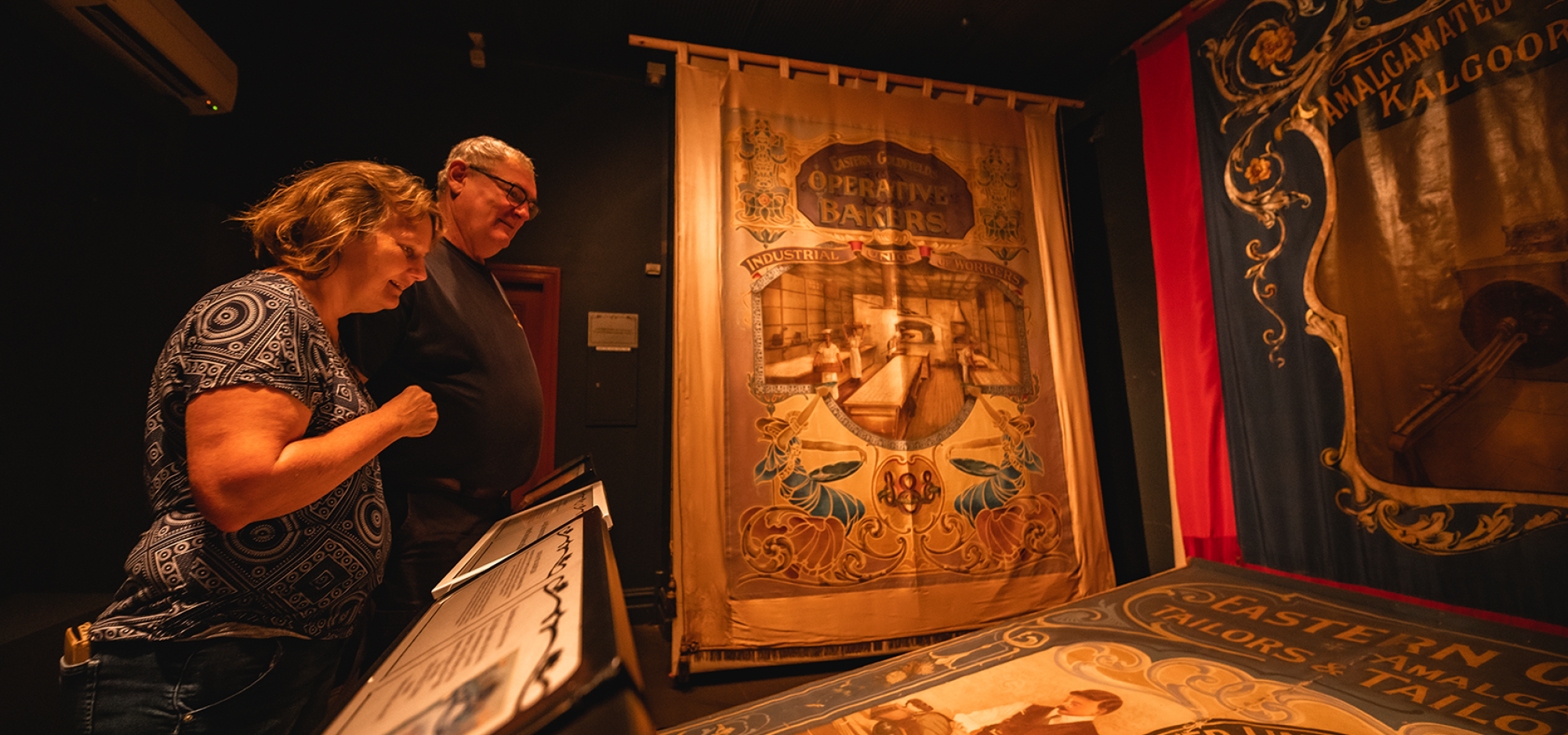 2 people observe a variety of banners on display in a dimly lit museum gallery