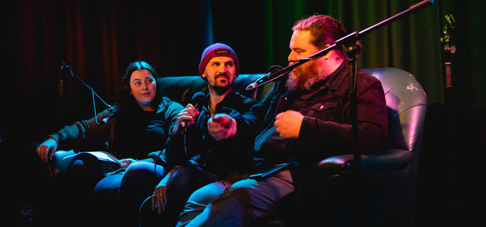 Three people sit in a dark room on a couch, speaking into microphones