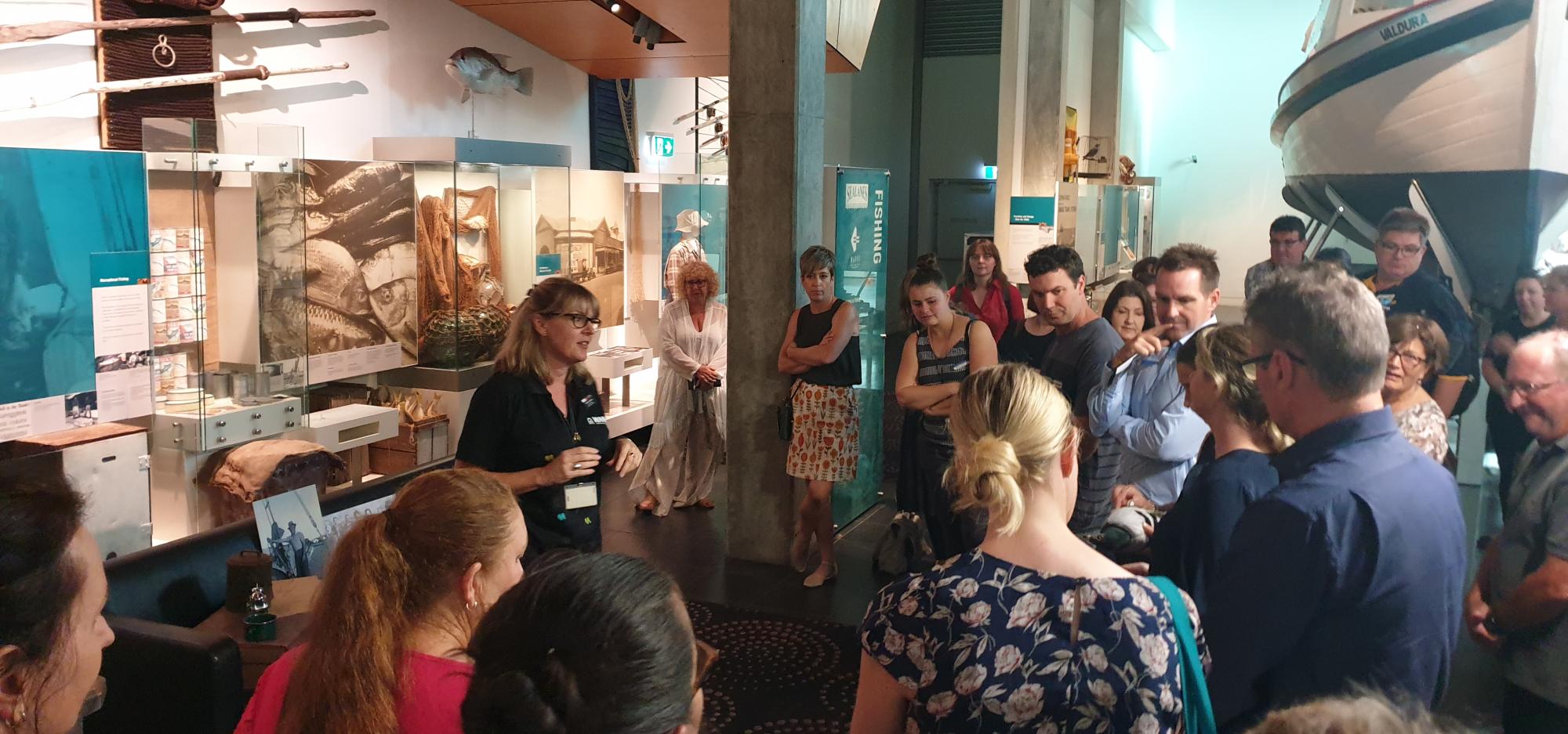 A group of teachers stand around a facilitator, who is giving a presentation in front of some showcases in a museum exhibition