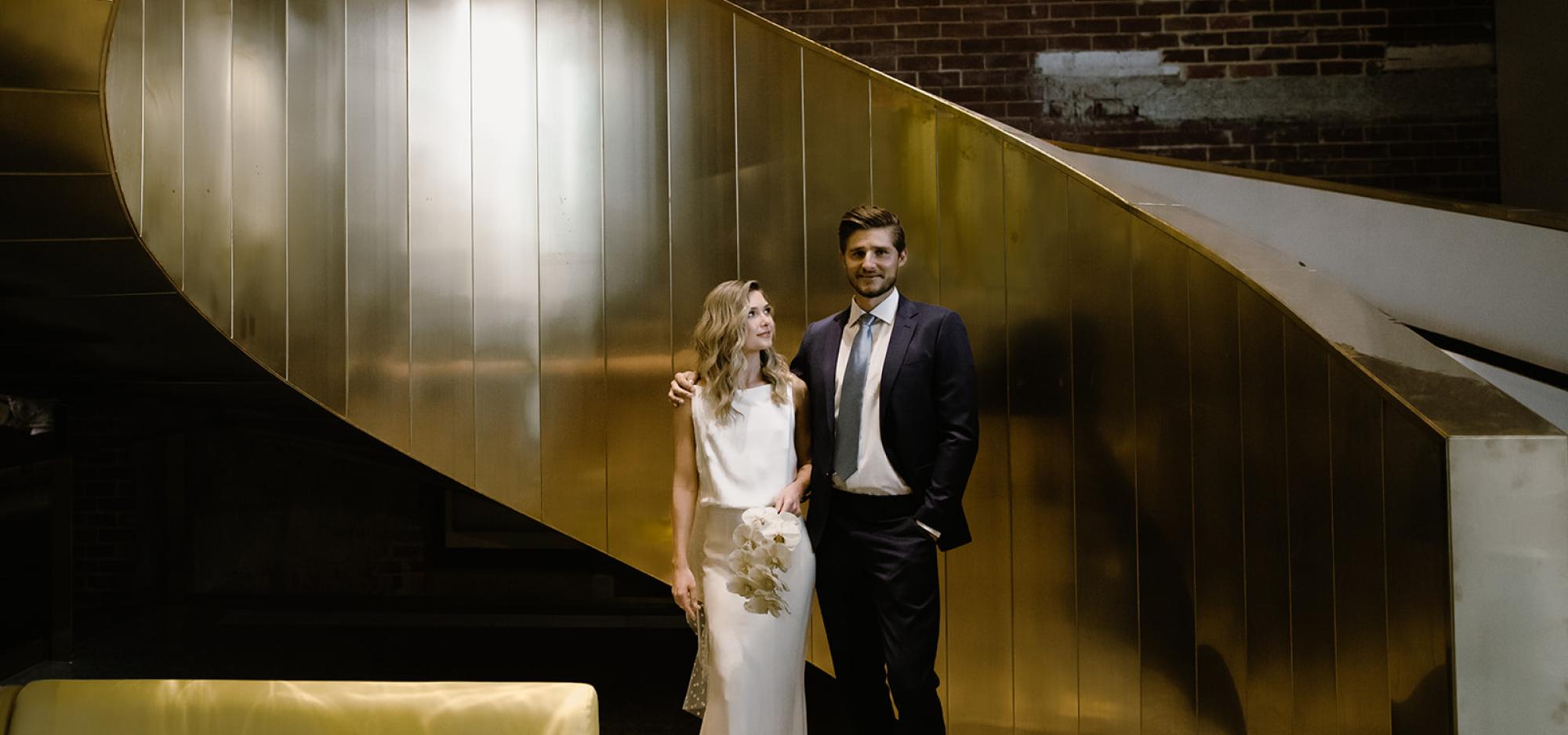 2 people dressed in a wedding dress and suit standing by a gold staircase