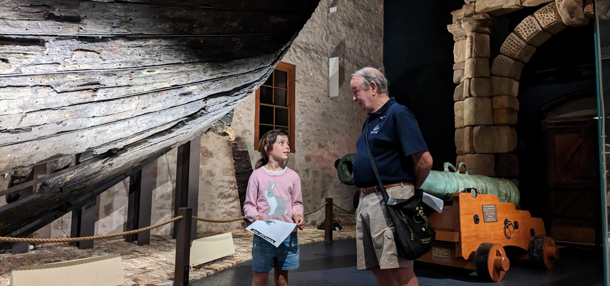 A mature male volunteer wearing a WA Museum badge stands in front of the hull of the Batavia shipwreck. A school-aged girl stands nearby, holding out a worksheet and discussing it with the volunteer.