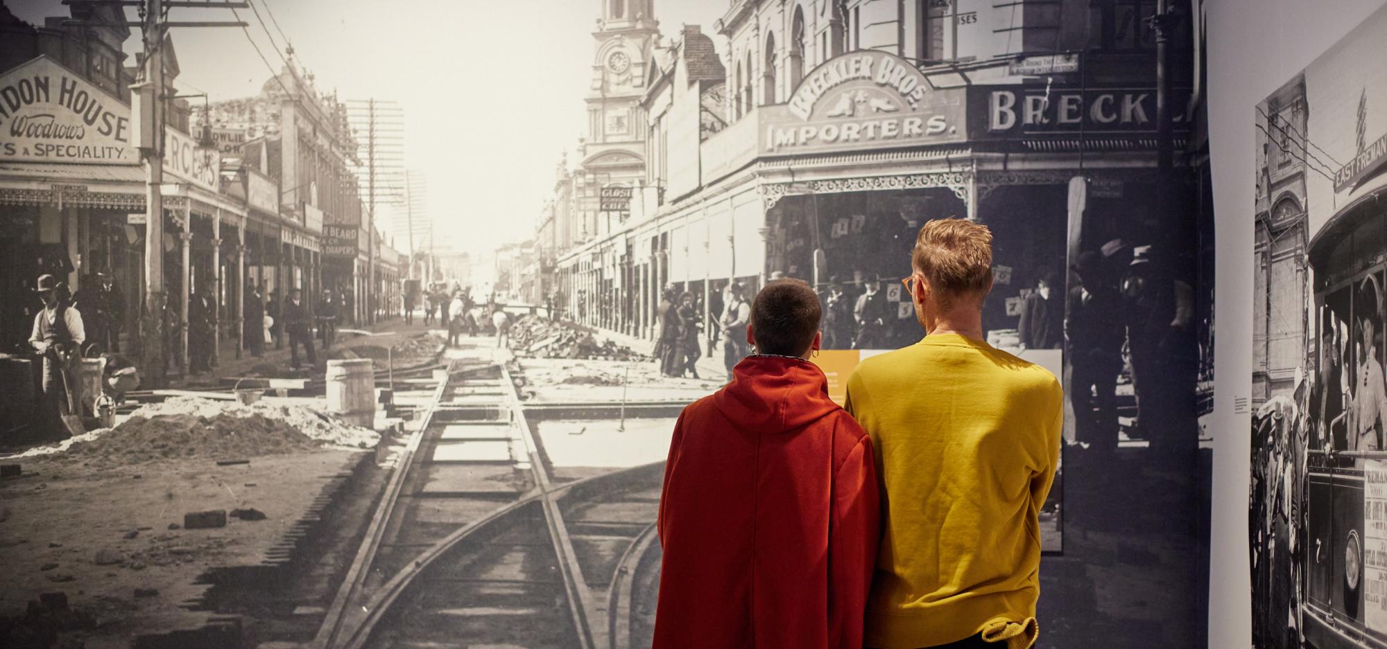 The image depicts two people with their backs to the camera staring at a large black and white historic photograph of workers building the Fremantle railway surrounded by old buildings.