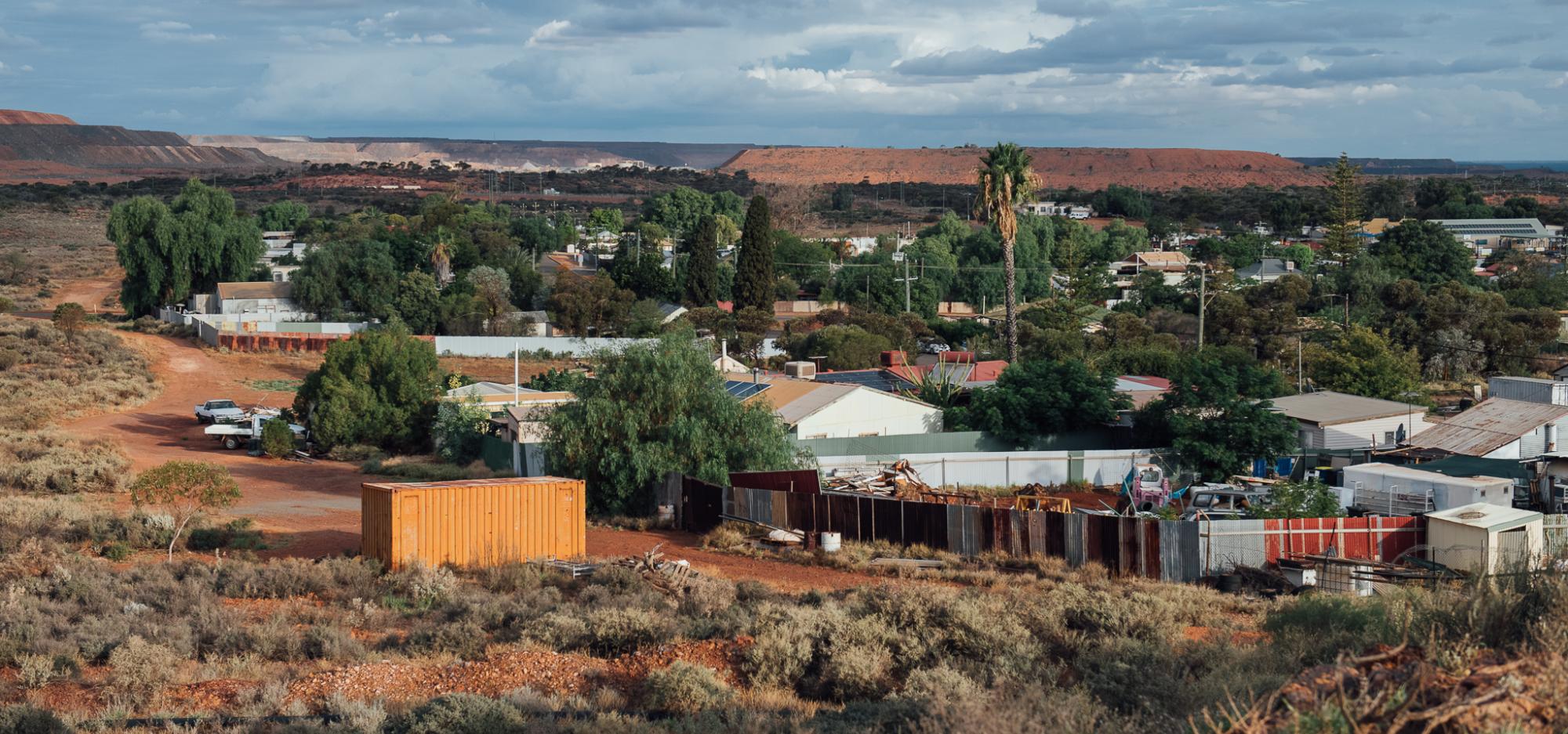A landscape image of Williamstown d epicting a series of tin roofs visible among red dirt and dark green trees. 