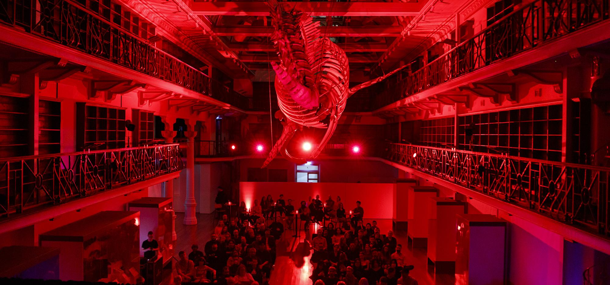 A large hall of people sitting in rows under a suspended whale skeleton in a heritage library building with empty book shelves. The whale and crowd is lit with a bright red light, giving the image a red glow