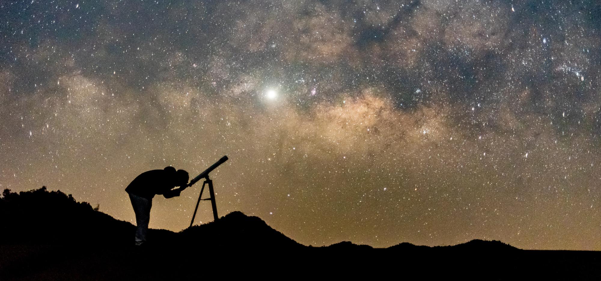 A silhouette of a person gazing through a telescope, in front of a vibrant night sky filled with stars, and deep blue and gold tones