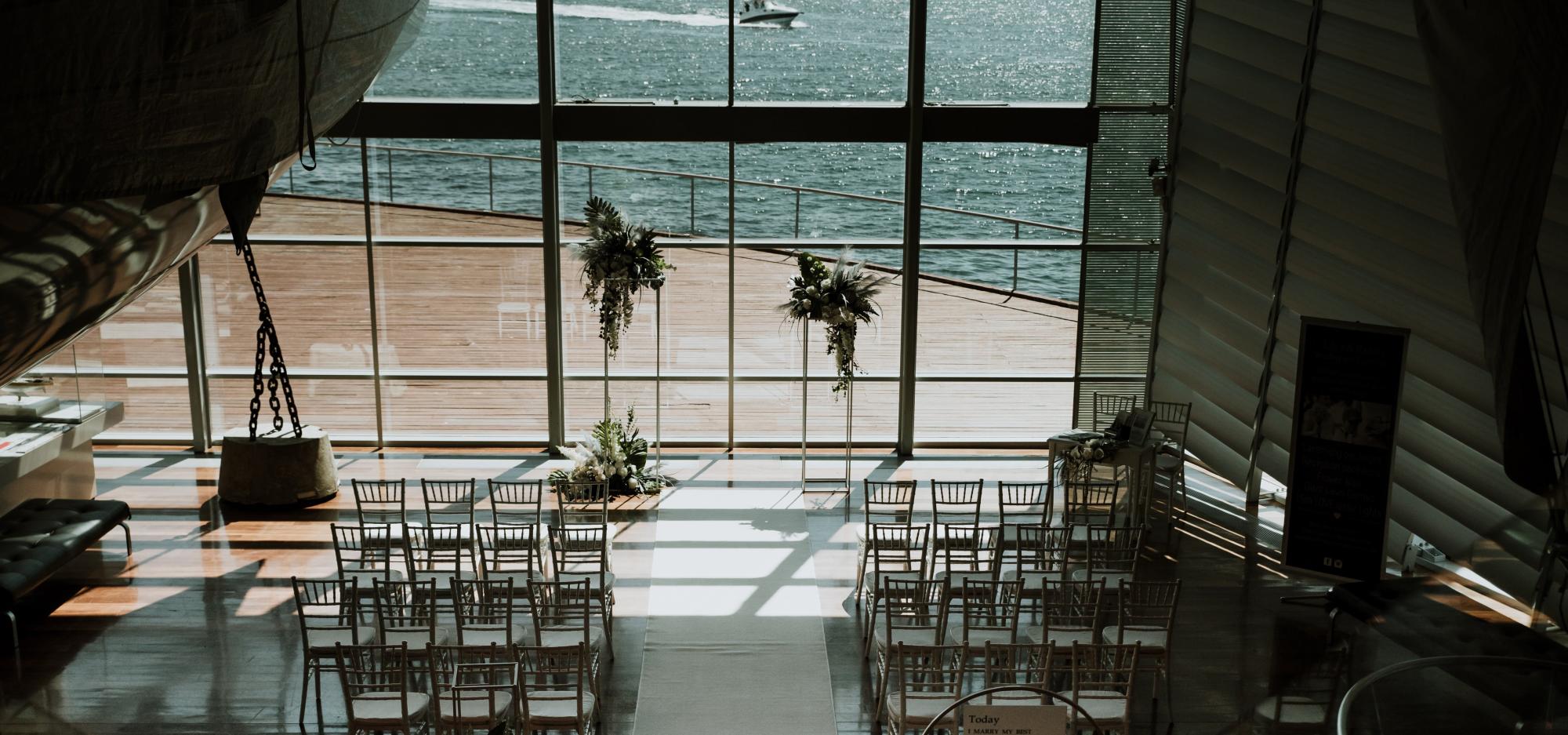 A high-angled photograph looking down a flight of narrow stairs towards two aisles of white chairs in front of a floral arch and large, brightly-lit window.