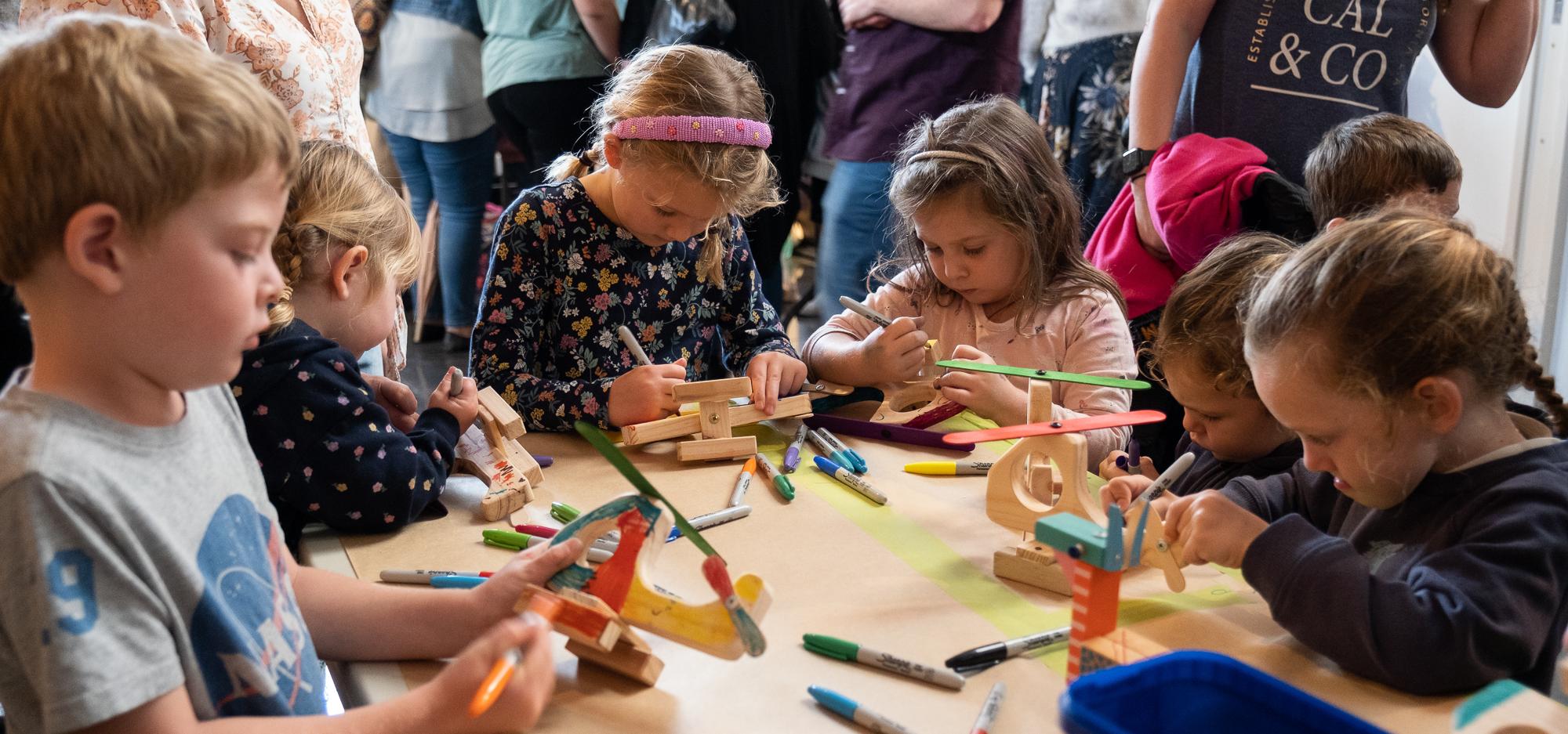 A group of children gathered around a table playing with various wooden arts and crafts items, and staring at their creations with intense concentration faces