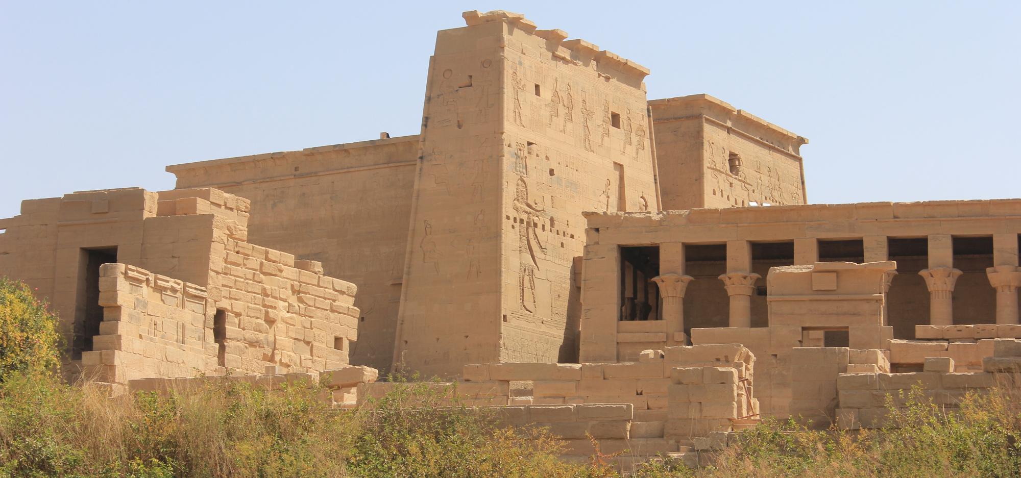 A land sandstone temple positioned in desert-like area with dry green shrubbery stands against a clear blue sky, engraved with large images of people in an Ancient Egyptian style