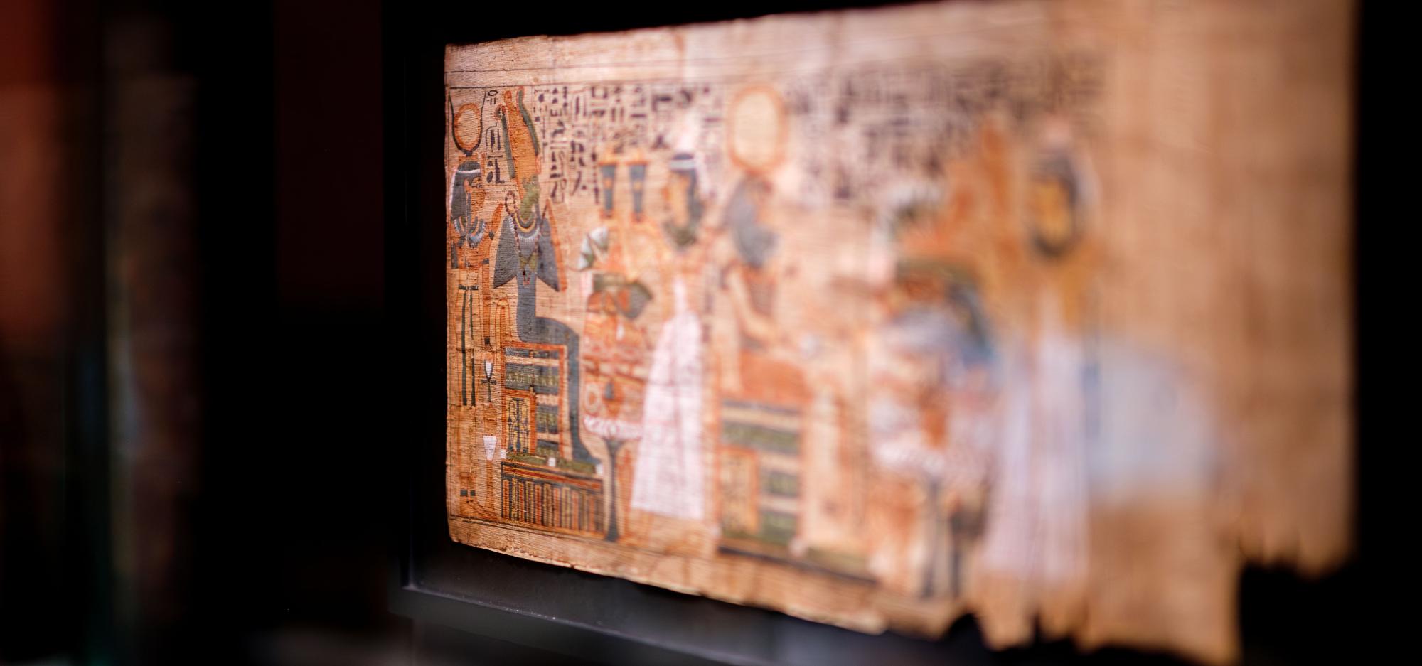 The Book of the Dead of Padikhonsu, a yellowed papyrus scroll with a colourful depiction of a god in mummifrom clutching a crook and flail while being attended by several Egyptian women surrounded by hieroglyphs